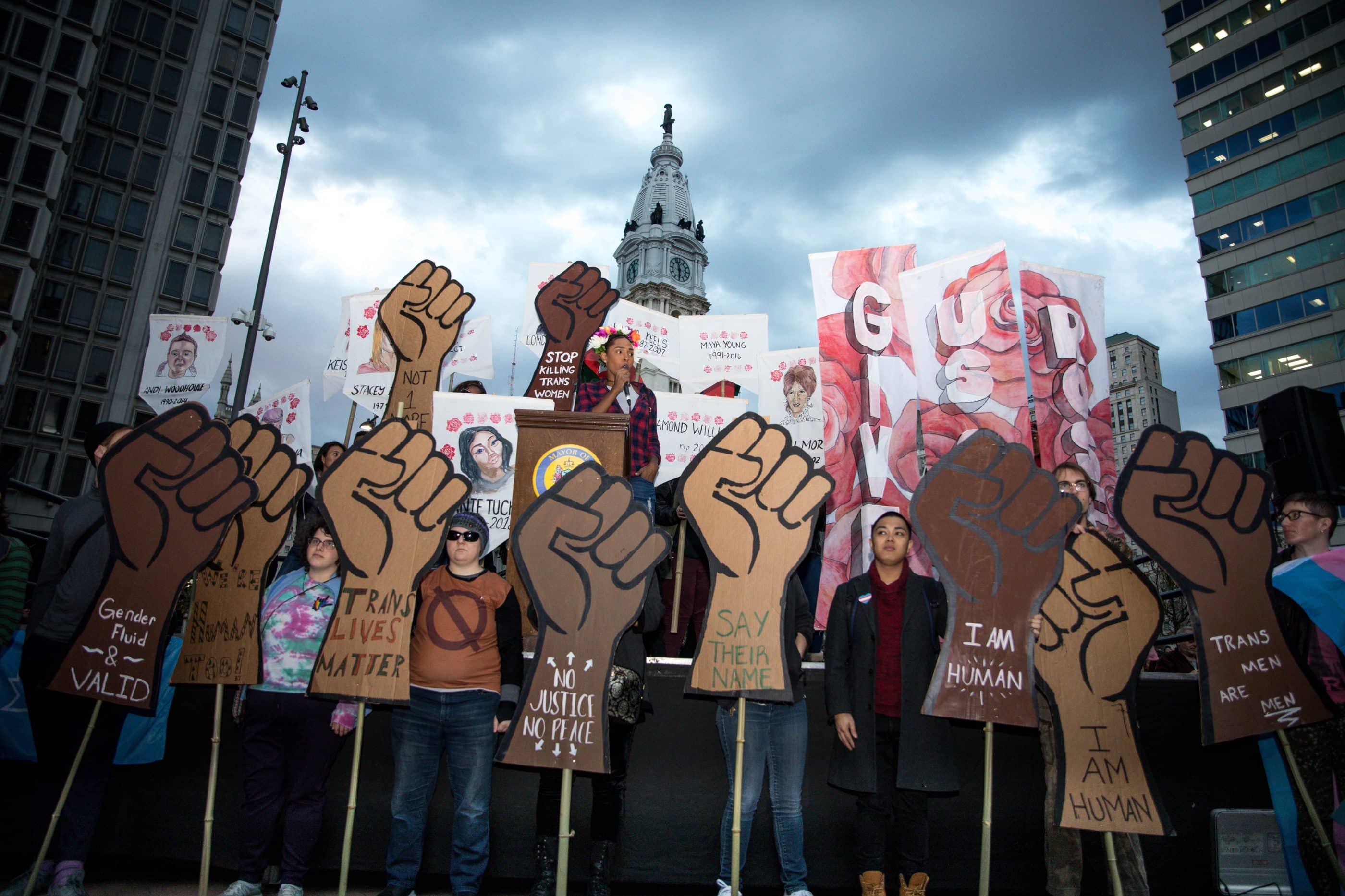 A group of people holding cutout fists stand in front of a building. The fists say "say their name," "I am human," "trans lives matter" and other messages. Behind the fists are banners with the faces and names of various people.