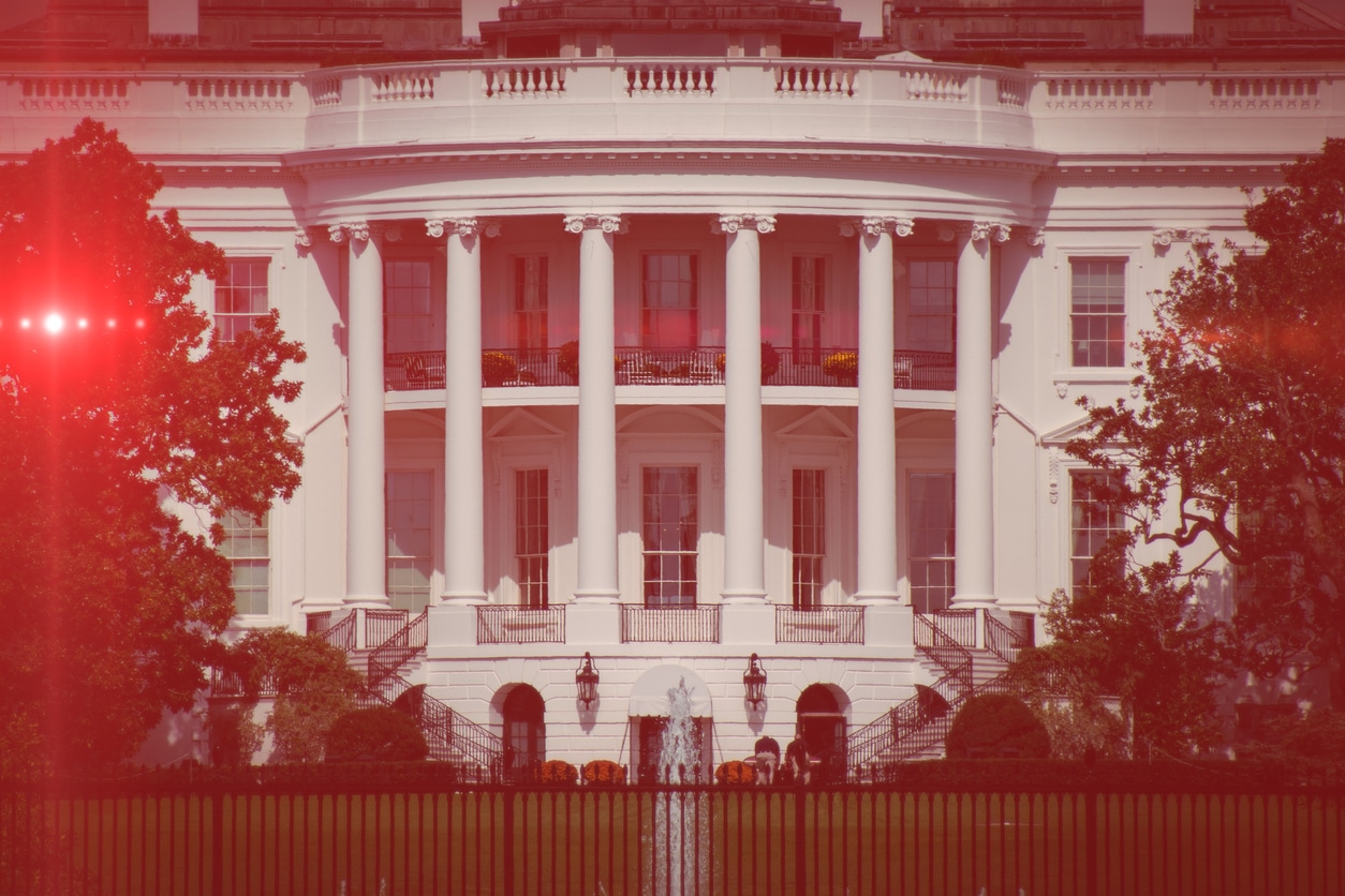 The portico of the White House overlooking the South Lawn. An imposing building with columns marking a two-story portico and a balcony on the second floor. Angled staircases on either side lead up to the first floor. On the lawn, trees in leaf frame the portico on either side. The photo has an eerie red tint.