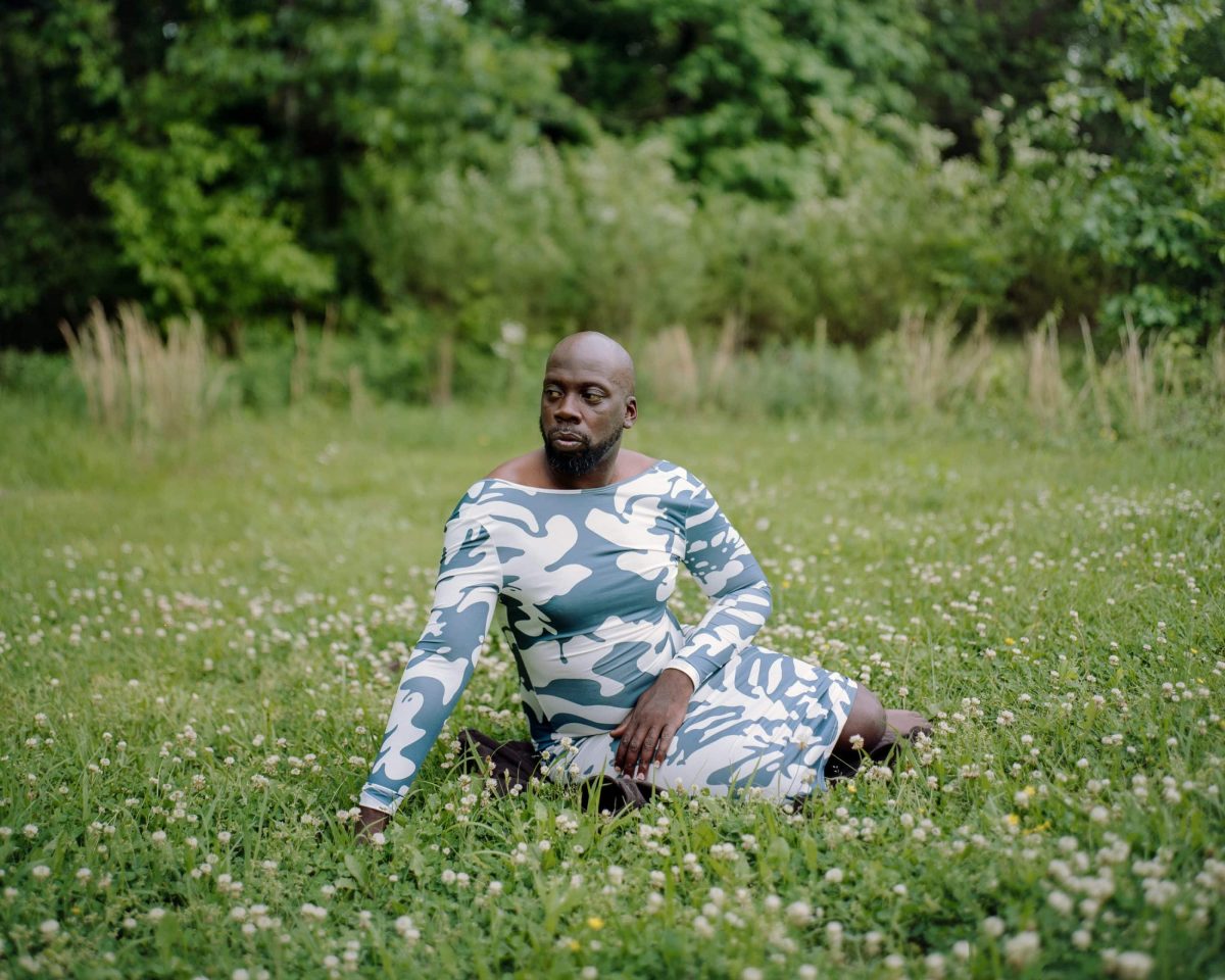 In an outdoor nature setting, a Black man sits in a field of grass and white flowers. The man is bald, has an extended goatee, and is looking to the left. He is wearing a blue and white long sleeve dress that is cut off at the ankles. One hand is resting in grass and the other on his lap.