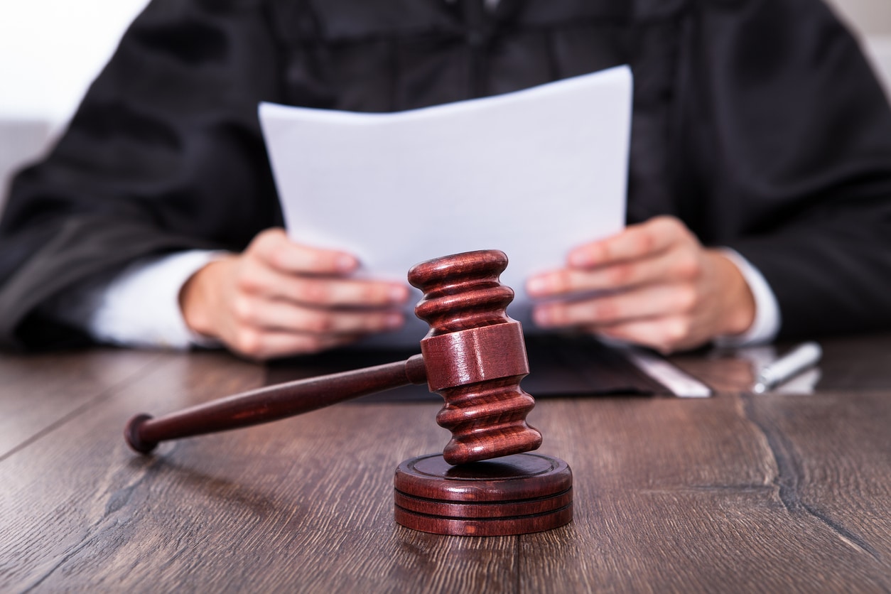 Close-up partial view of white male judge in black legal robe at a wooden table holding papers. In front, in focus, is a wooden gavel.