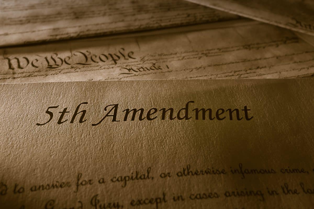 Close view of reproduction of the 5th Amendment with U.S. Constitution behind it. Low angled light shows the texture of paper. Text of amendment that is visible reads "to answer for a capital, or otherwise infamous crime,/(next line) except in cases arising in the." The document in the background shows only the preamble and "Article I."