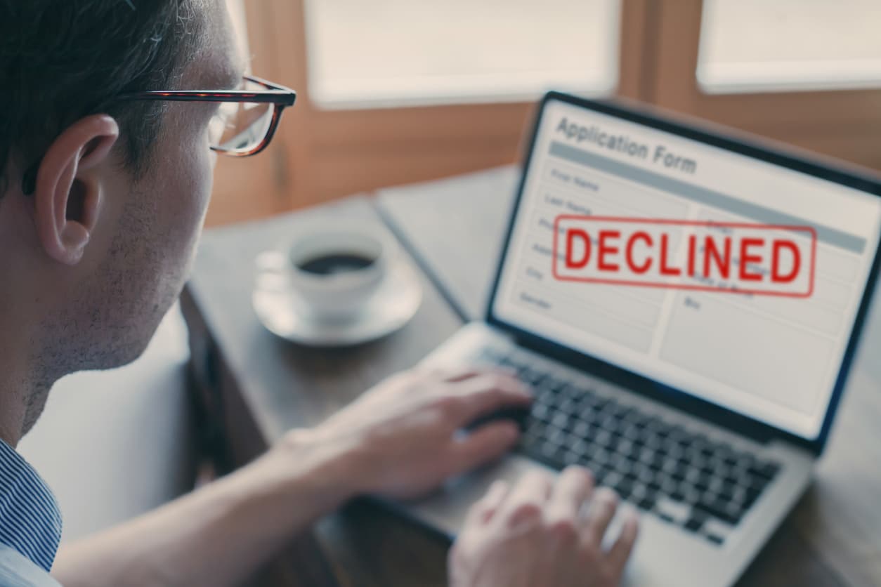 A person wearing glasses sits at a table with his hands on the keyboard of a laptop. On the screen is a denial for an application