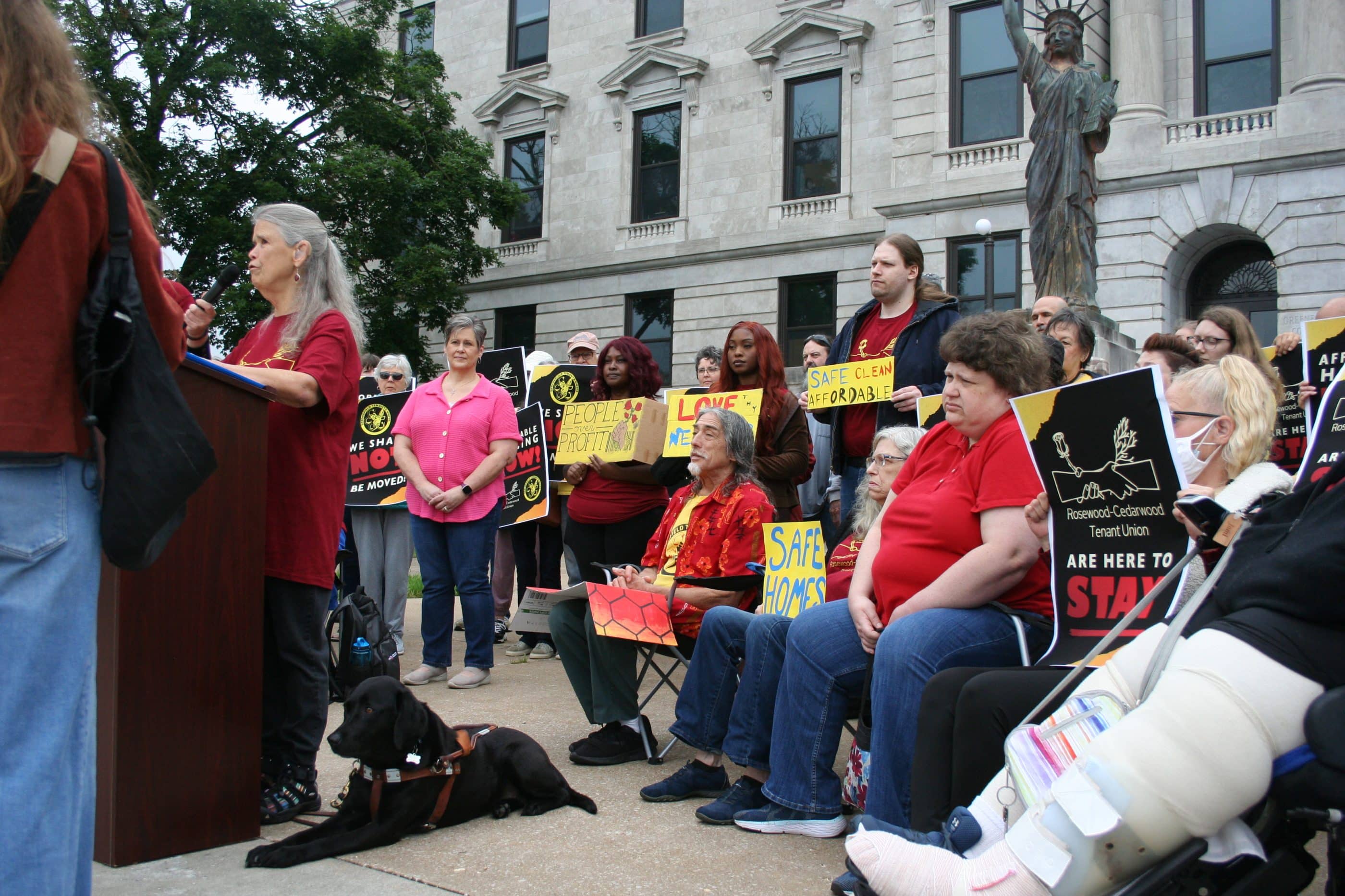 A light-skinned woman with long gray hair speaks at a podium at an outdoor rally. On the ground to her left is a black dog in a service dog harness. Behind her are various tenants holding signs.