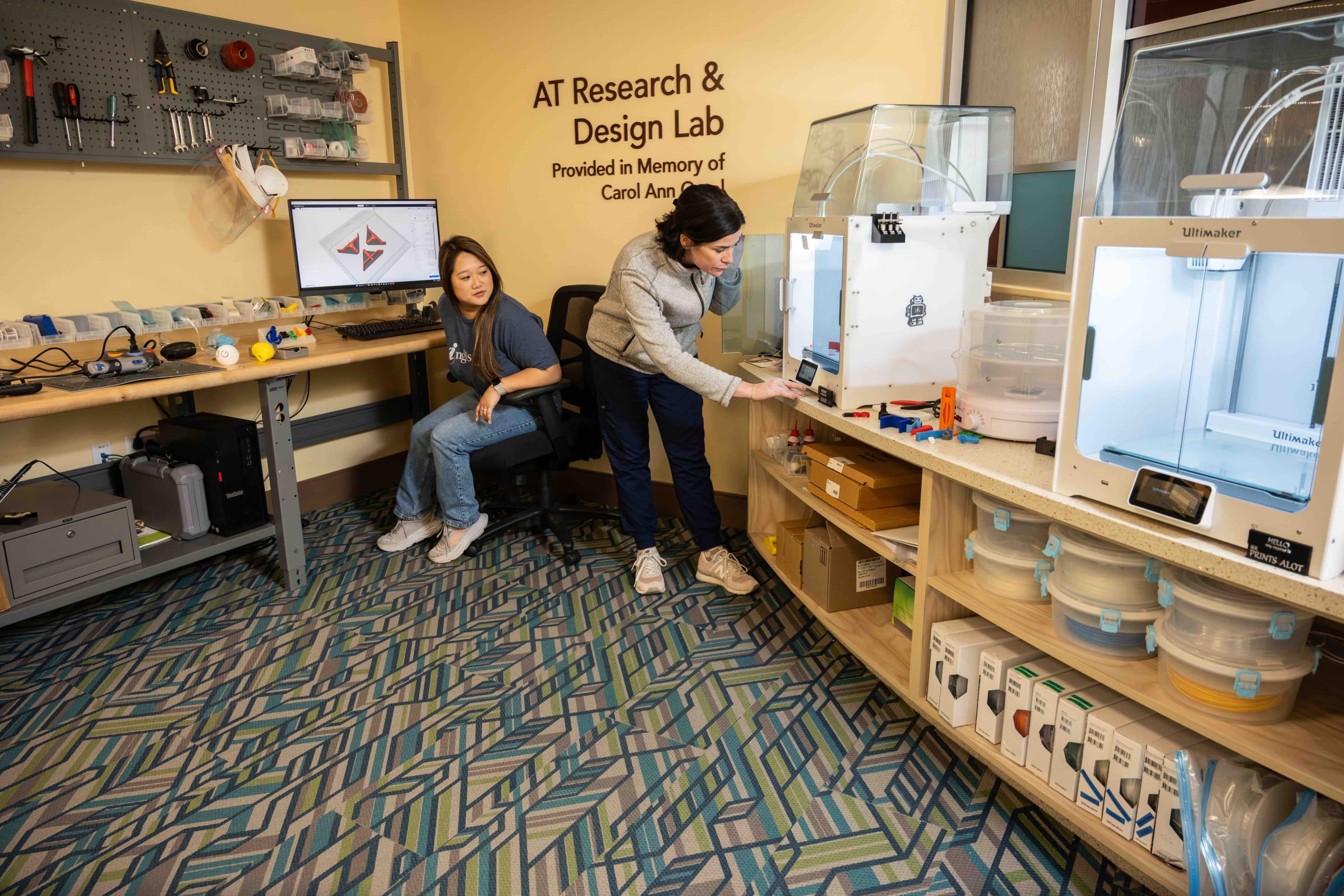 Two young women in casual clothes in a modern office. On the far wall, a sign says "AT Research & Design Lab/Provided in Memory of Carol Ann"—with the rest of the name obscured by one of the women. Pegboard on the adjoining wall holds various tools, and the shelves contain boxes and software.