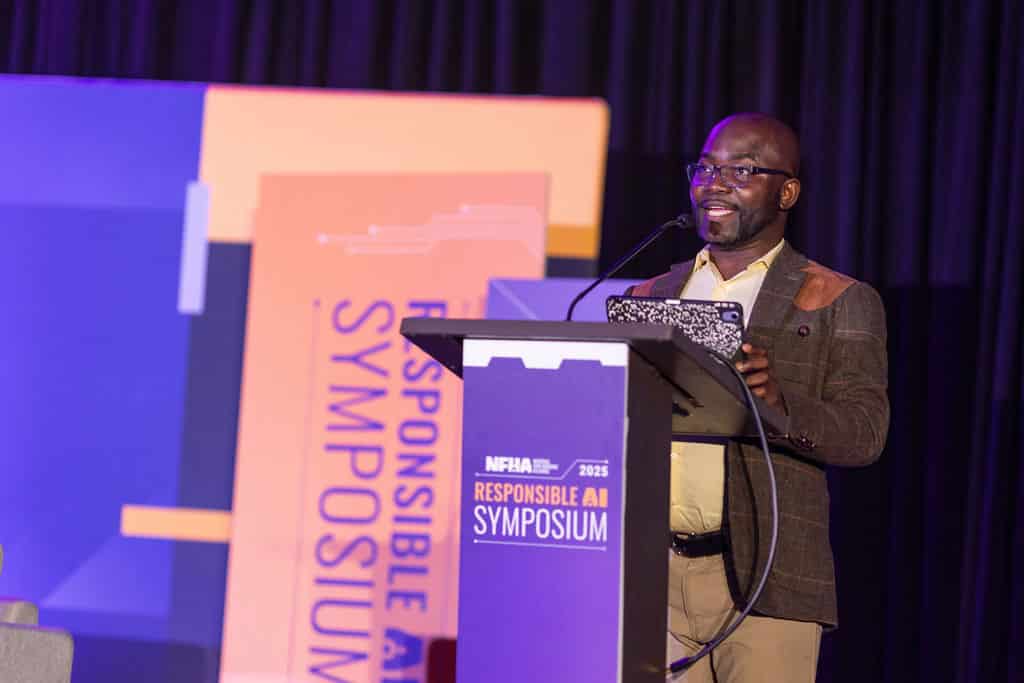 A black man, who is wearing glasses and holding a black and white tablet, speaks from a podium. He is wearing a yellow shirt and a brown striped jacket. In front the podium is a purple sign that reads, Responsible AI Symposium.