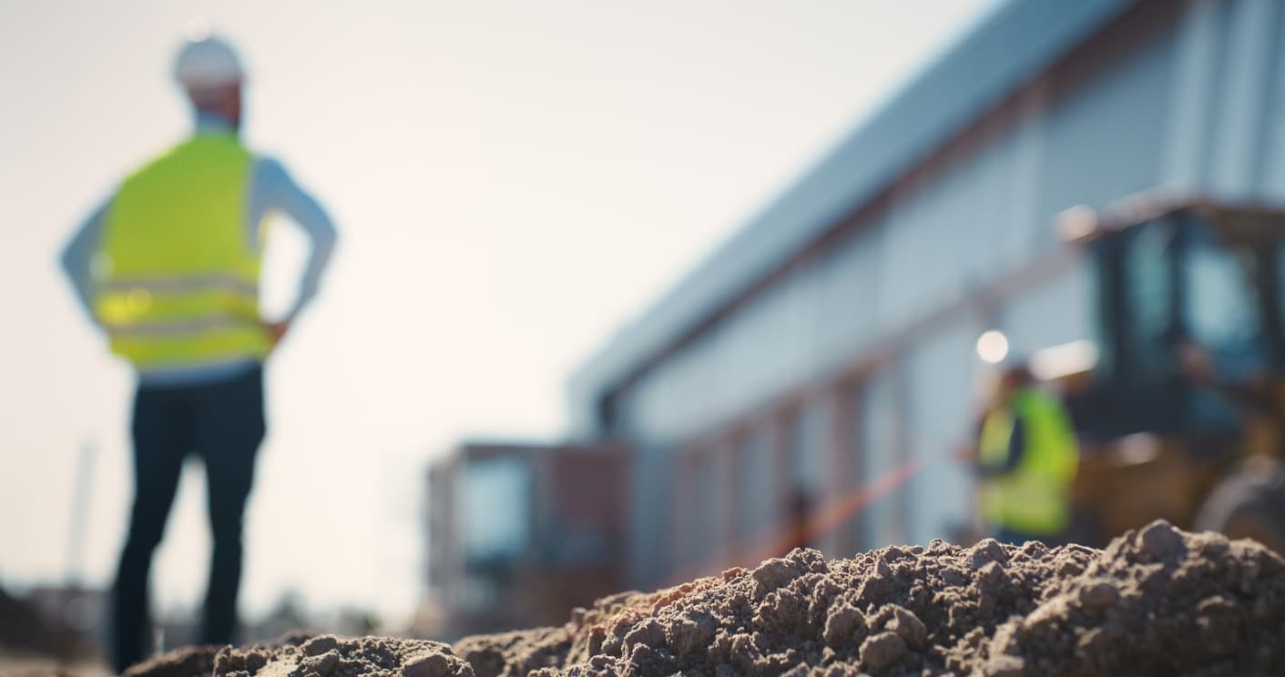 Blurry view of man in hardhat and safety vest with back to camera, facing partially built building. In the near foreground, in focus, is loose soil from recent digging.