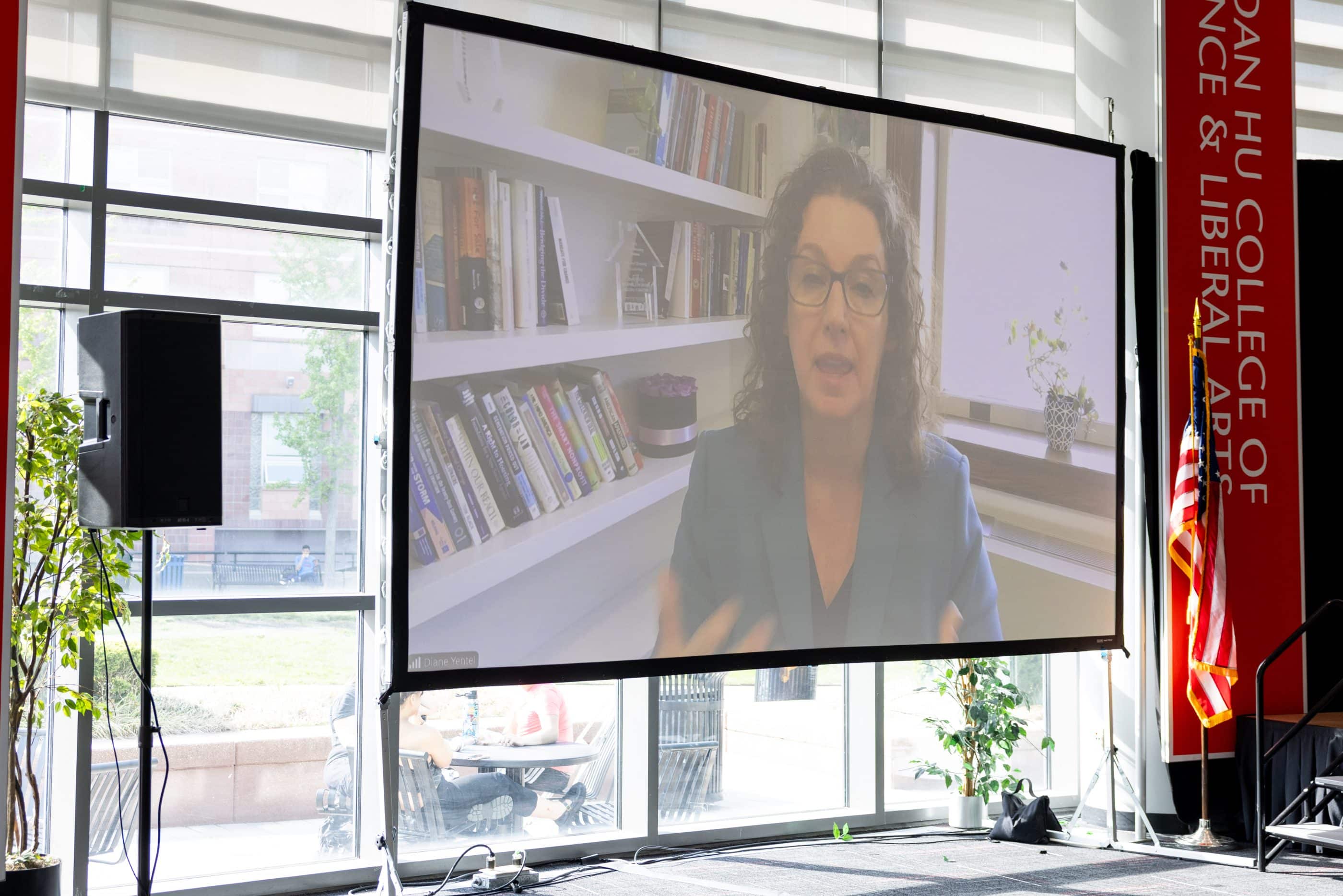 Large screen in a sunlit conference room shows a white woman with curly hair and wearing glasses speaking remotely. Audience is not in the frame. Behind her is a bookshelf and sunny window.