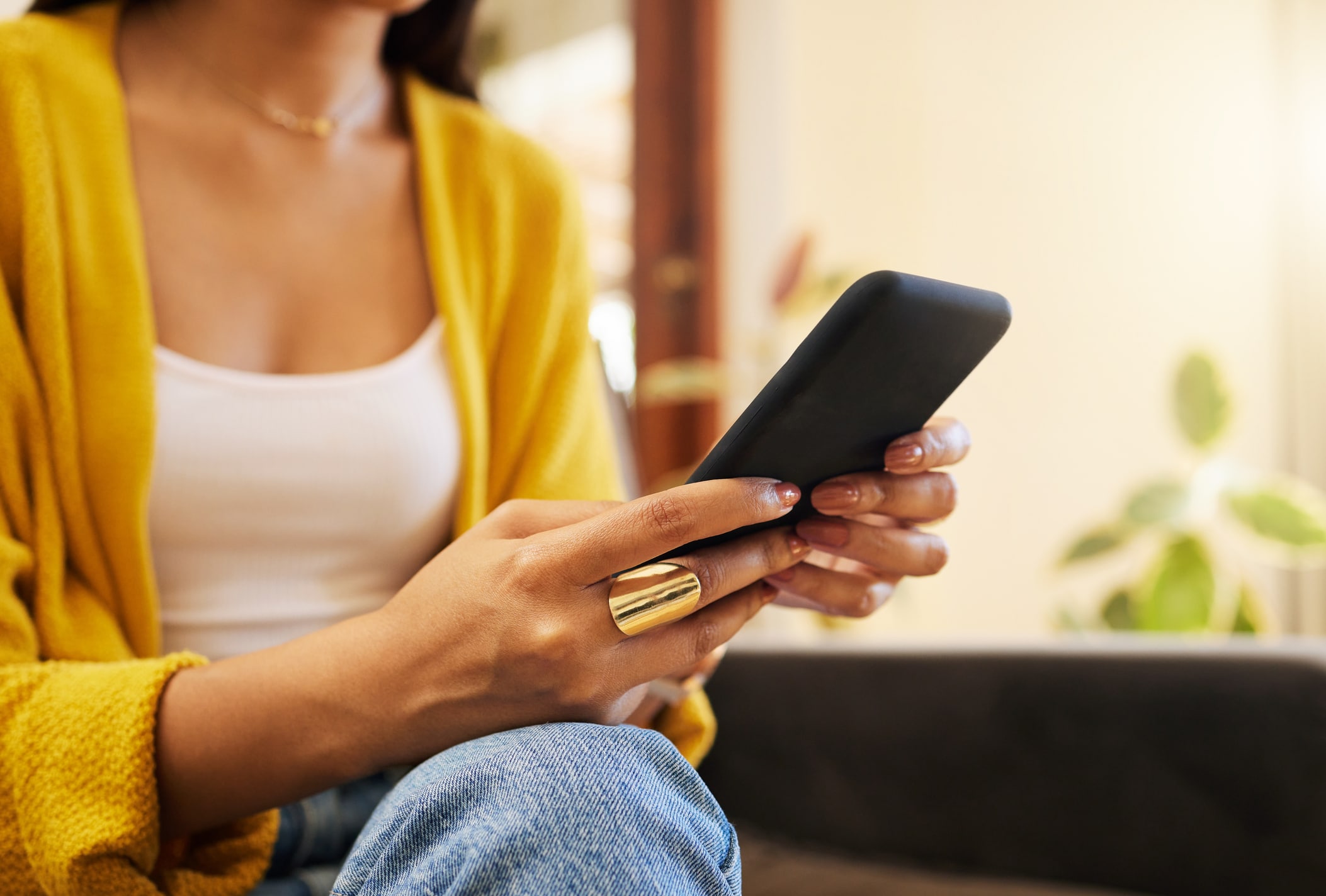 Closeup of a young woman in a yellow sweater and white shirt using her smartphone while sitting on a sofa in a bright living room.