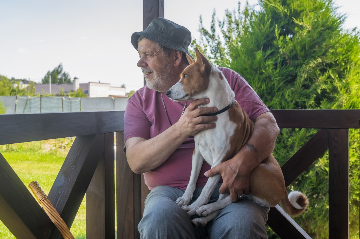 A white-whiskered white man in a blue bucket hat, pinkish-red T-shirt, and dungarees sits on a porch holding a brown-and-white dog. They are both looking off to the left at something out of frame. Behind them to the right is an evergreen, possibly juniper, tree, and way off to the left, a wire fence and some nondescript buildings.