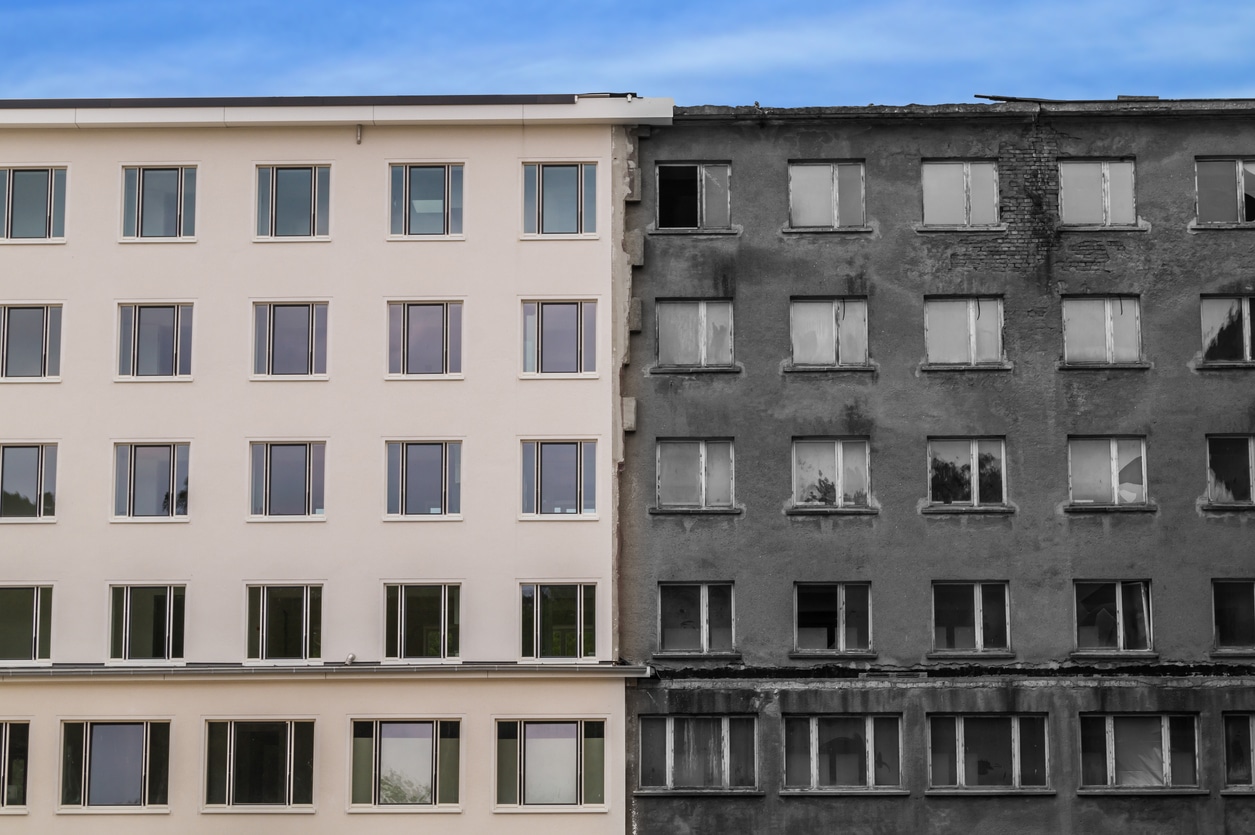 Two very similar apartment buildings side by side fill the frame, with a strip of blue sky above. The one on the left has been renovated and painted a putty color, and looks fresh and clean. The one on the right looks old and dirty and has missing windows and broken masonry.