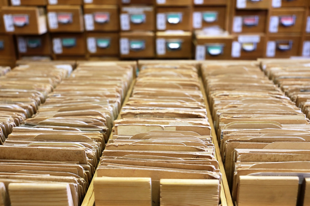 Close view of five rows of open shallow drawers filled with yellowing, dog-eared paper file cards and manila divider tabs. Beyond the card boxes and somewhat blurry is what looks like the front of an old-school library card catalogue, with closed drawers.