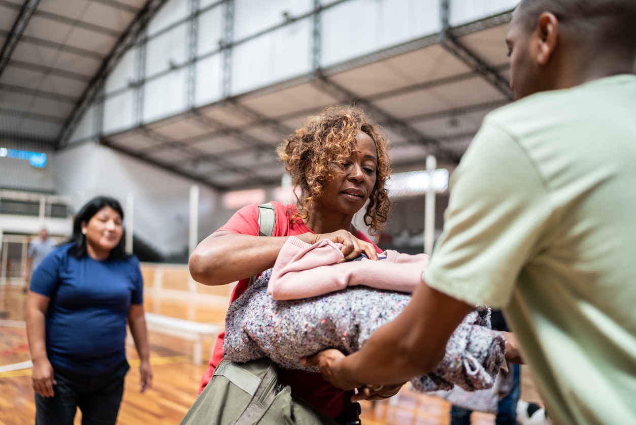 In a large, high-ceilinged room like a community center, a Black woman in a reddish shirt is handing bedding (gray and pink blankets or covers) to a Black man in a light green T-shirt. Behind them is a second woman, dark-haired and wearing a blue shirt.
