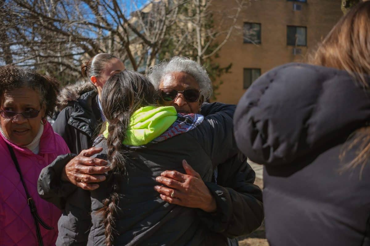 Two women hugging, surrounded by other people. The setting is outside a multistory apartment building.