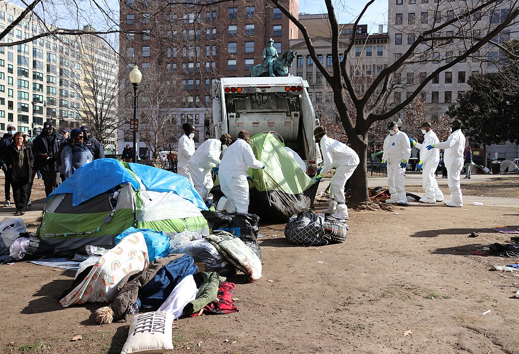 In a city square, perhaps a park, four workers in white protective jumpsuits and wearing masks lift a tent into the back of a garbage truck, while three others look on. Nearby are other tents and jumbled personal belongings, blankets, and clothes. In the foreground, a throw pillow printed with the words "But first, coffee" lies in the dirt.