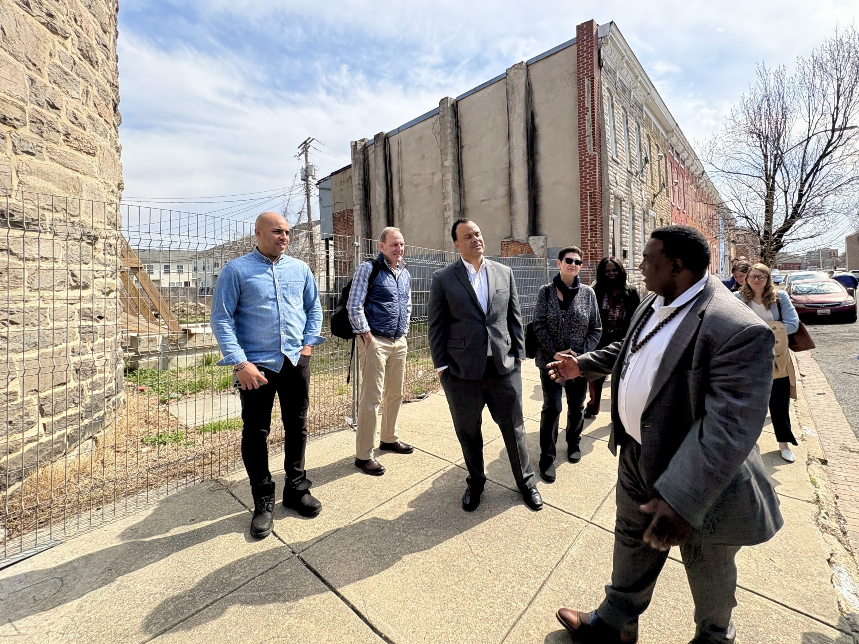 Outside a city street, a group of people—men and women—listen to a black man who is giving a tour of a new development. The man is wearing a gray jacket and paints, a white shirt, and a necklace.