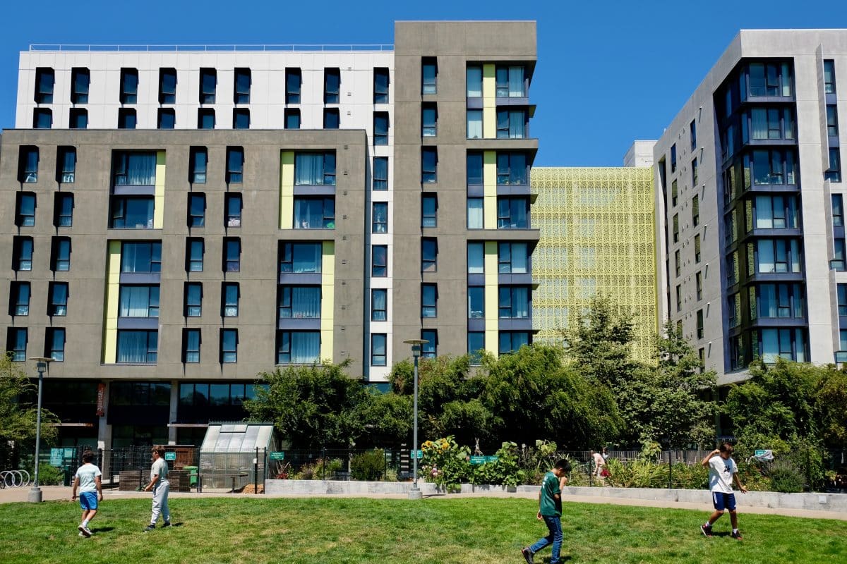 Multi-story apartment buildings in shades of brown, beige, and eggshell, with ground-floor space that's not residential. On a lawn in front of one of the buildings, four boys are walking or running around. The sky is blue.