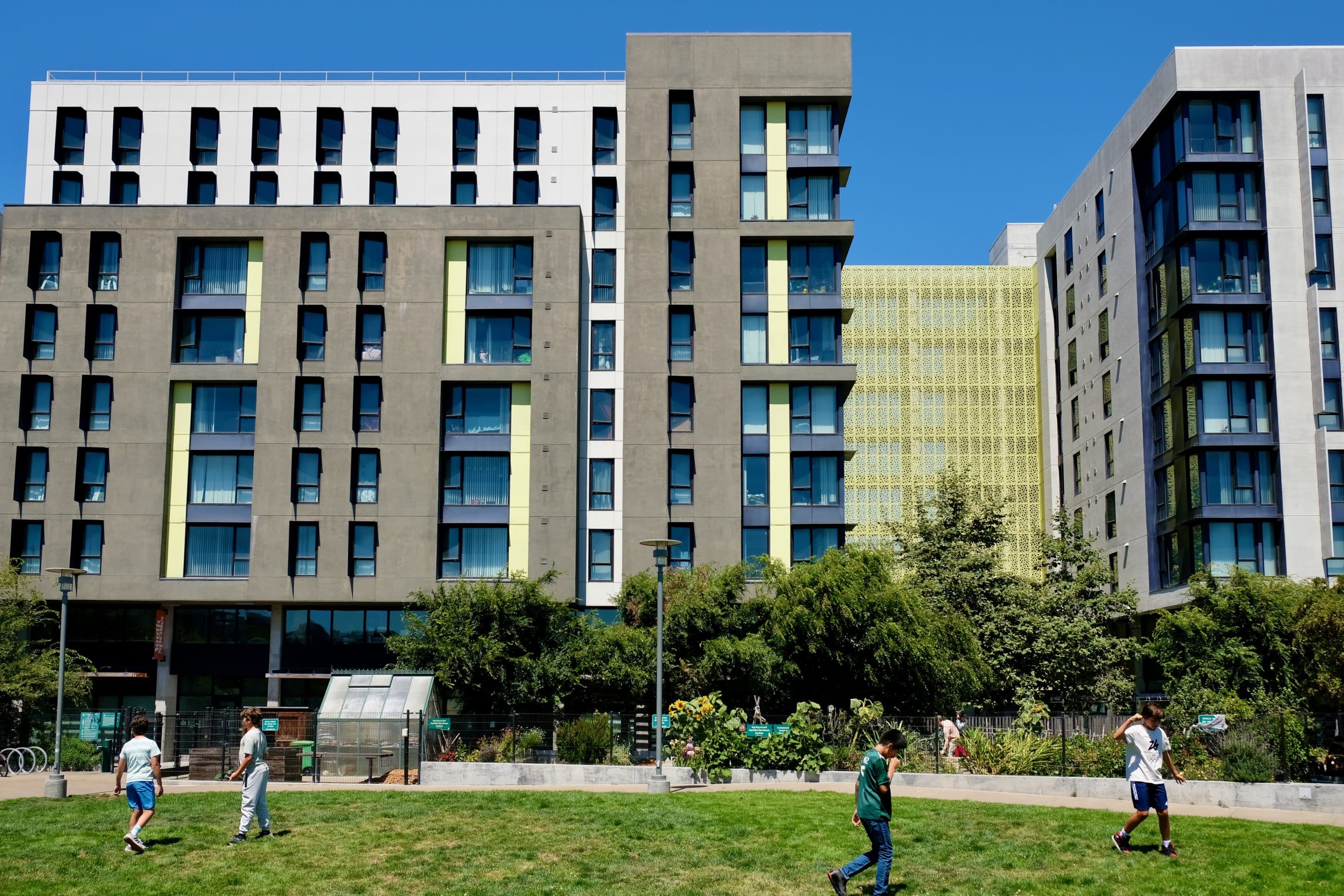 Multi-story apartment buildings in shades of brown, beige, and eggshell, with ground-floor space that's not residential. On a lawn in front of one of the buildings, four boys are walking or running around. The sky is blue.