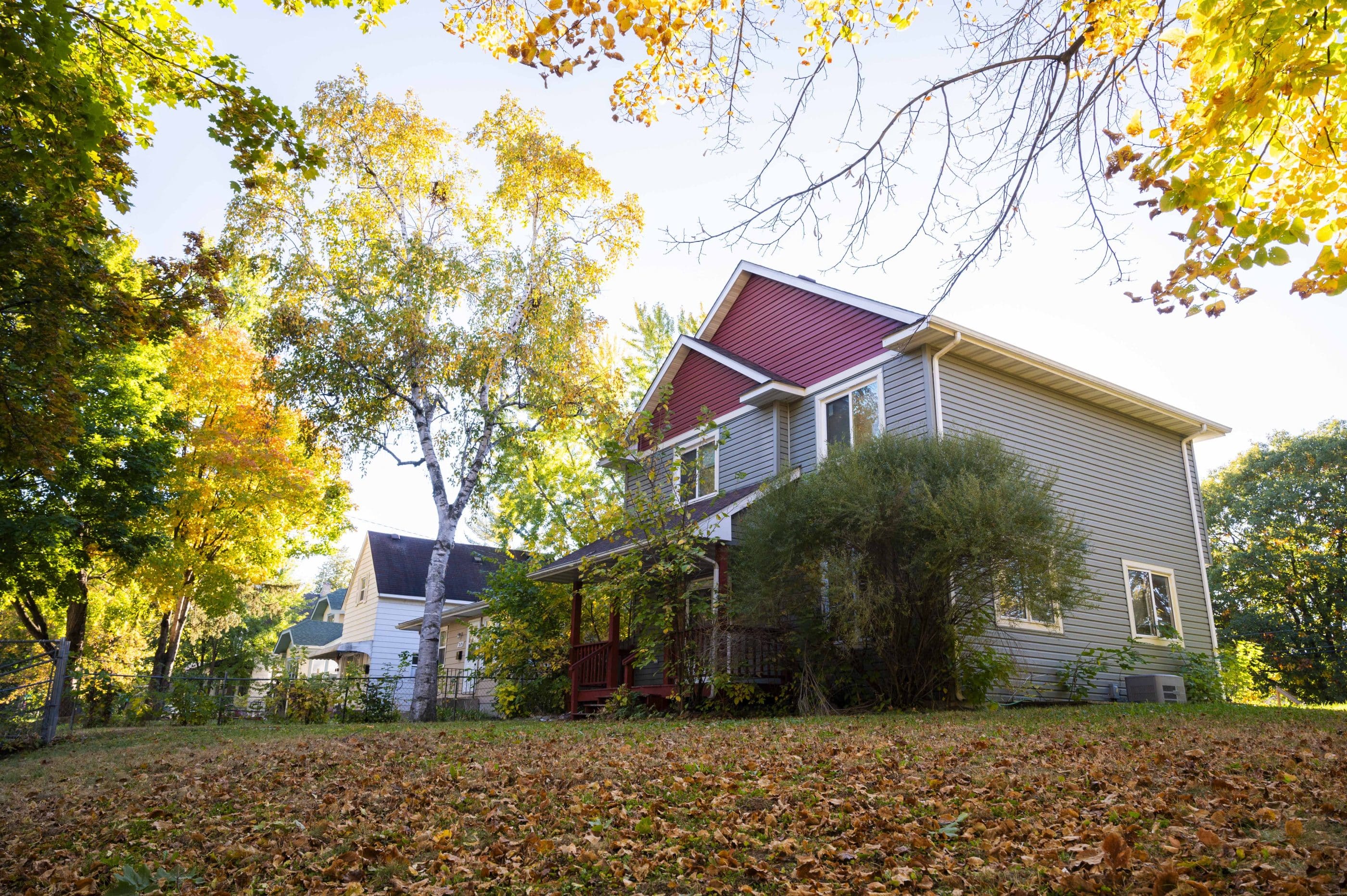 A pitched-roof house on a grassy plot covered with autumn leaves is mostly gray with dark red paint on the attic clapboard. Shrubs partly hide the ground floor windows and porch. Three or four houses are partly visible beyond the main one, and colorful trees fill the front yards.