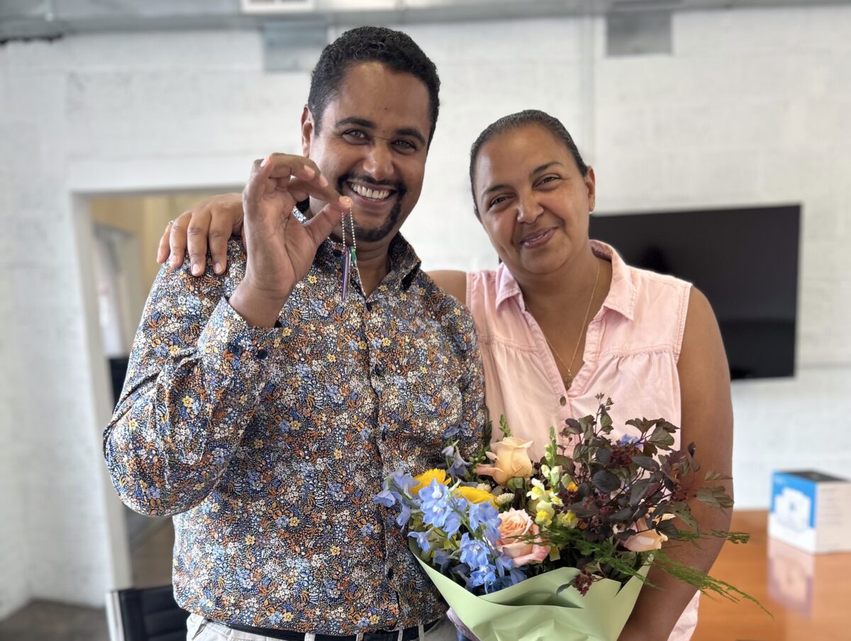 A man and woman with light brown skin stand close together, smiling at the camera. He has dark hair and goatee and wears a patterned shirt, and he's holding a little chain with keys on it. Her arm is around his shoulder and she holds a colorful bouquet. Her dark hair is pulled back and he's wearing a peach sleeveless blouse.