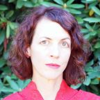 A white woman with short, red-brown hair, shown from the shoulders up in front of greenery. She is wearing red.