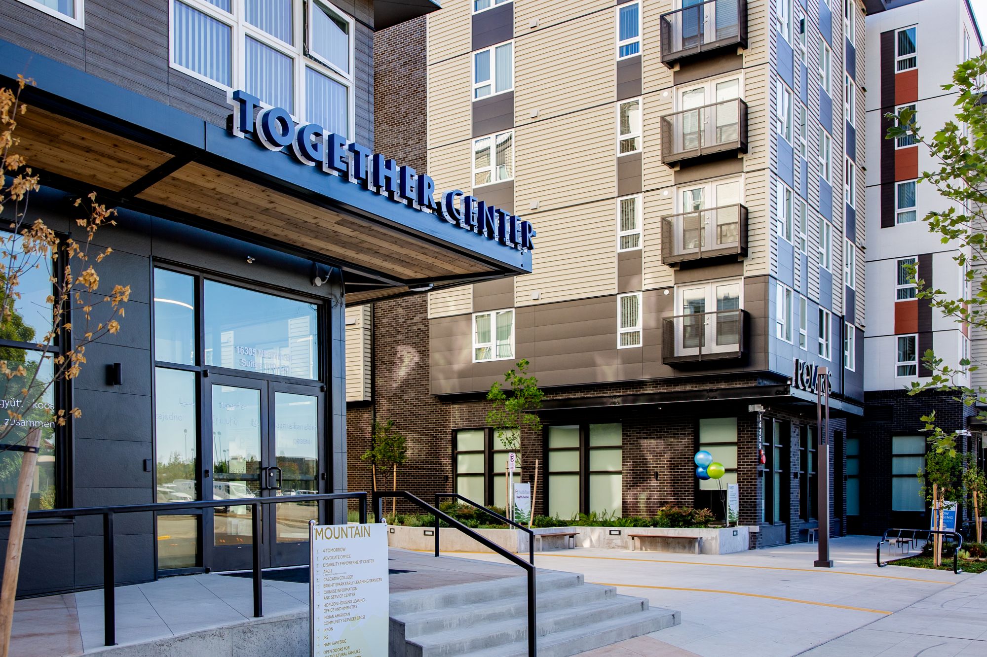 Angled view of front steps and front door of a ground-floor space (of a multi-story building). Over the door is an all-caps sign reading Together Center. Beyond the building is a similar-looking one, showing four floors above the ground floor. There are no people in the photo.