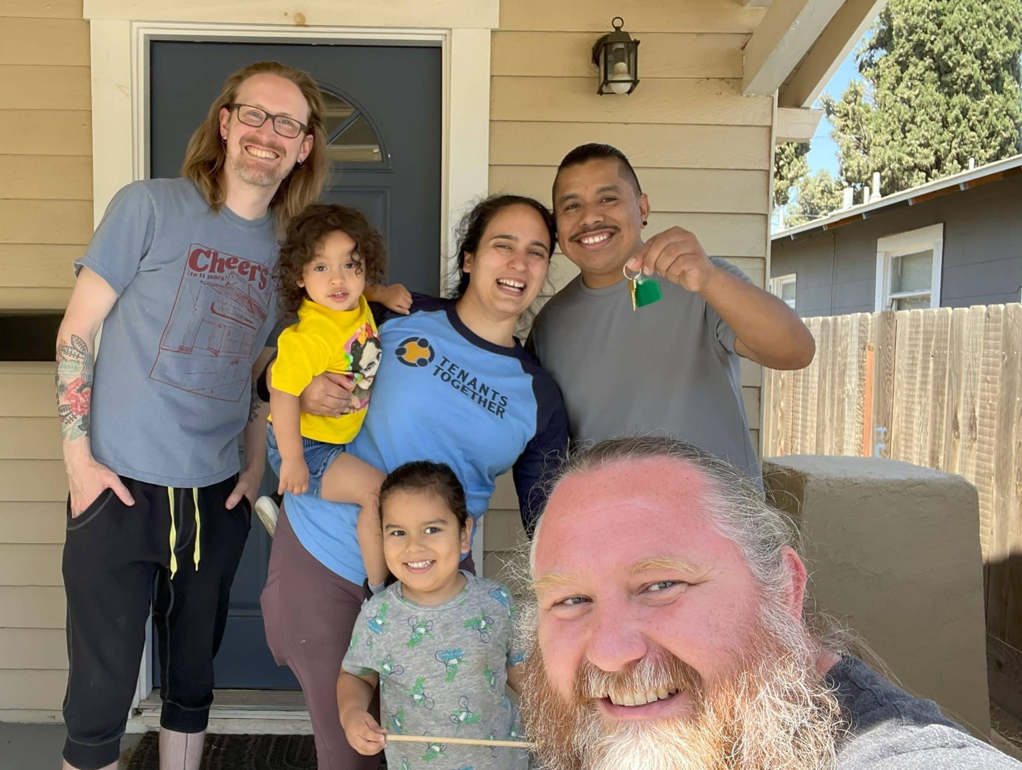 On the front porch of a tan-painted house are three adults and two children, with a fourth adult in the foreground, presumably holding a camera phone. All are smiling broadly, and the man at right holds up a keychain with a house key on it.