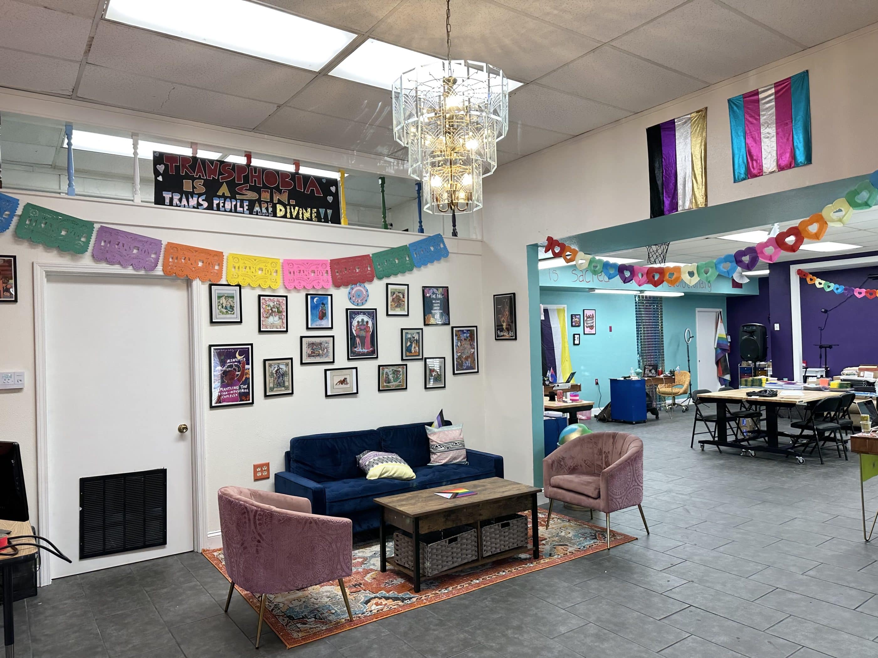 Two large rooms with high ceilings, decorated with many framed pictures as well as paper chains and banners. In the near room, a small sofa and two chairs surround a coffee table. Beyond, in the larger room are several tables and chairs, and signs of much creative activity, though no people are in the photo.