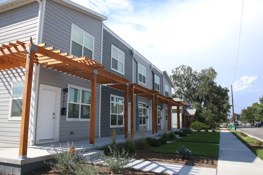Seen from one end, a row of attached houses in shades of gray line a suburban-looking street. Small grassy front yards separate them from the sidewalk.