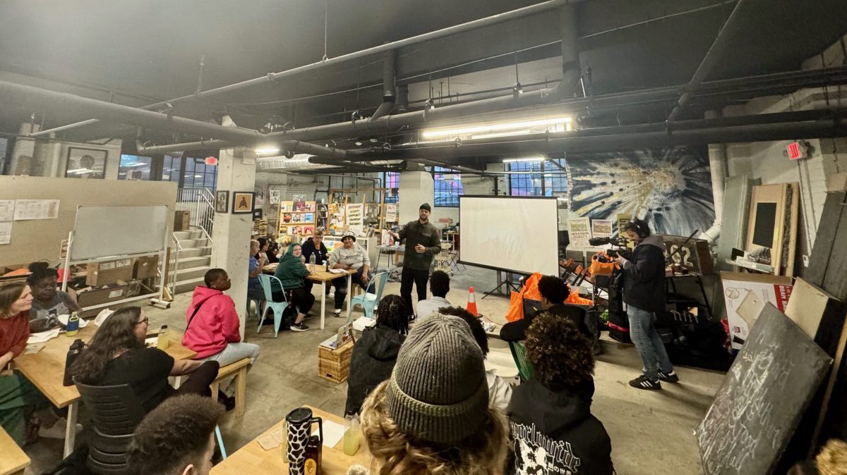 In a large space with visible overhead pipes and ducts, several people of mixed ages and skin tones seated at tables are listening to a man speaking and gesturing. At right is a man with a video camera pointed at the speaker. The room is crowded with posters and pictures on walls and columns.