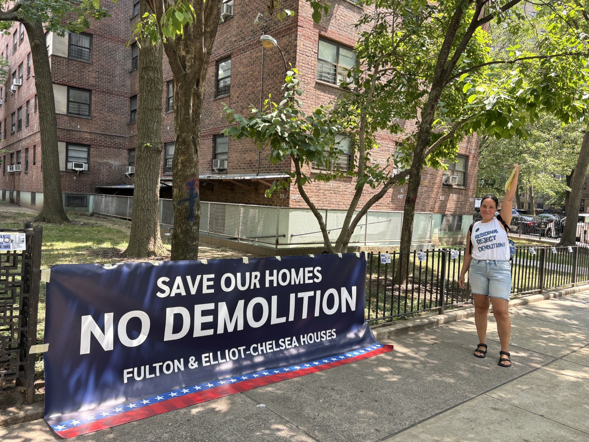 A woman with pulled-back dark hair and wearing cut-off shorts and a sleeveless shirt that reads "Residents/reject/demolition!" stands on the sidewalk with one arm raised high. Near her, attached to a short metal railing, is a large blue banner with white lettering that reads "Save our homes/No Demolition/Fulton & Elliot-Chelsea Houses." Inside the fence, across a small lawn, is a brick multistory apartment building, three stories of which are within the frame.