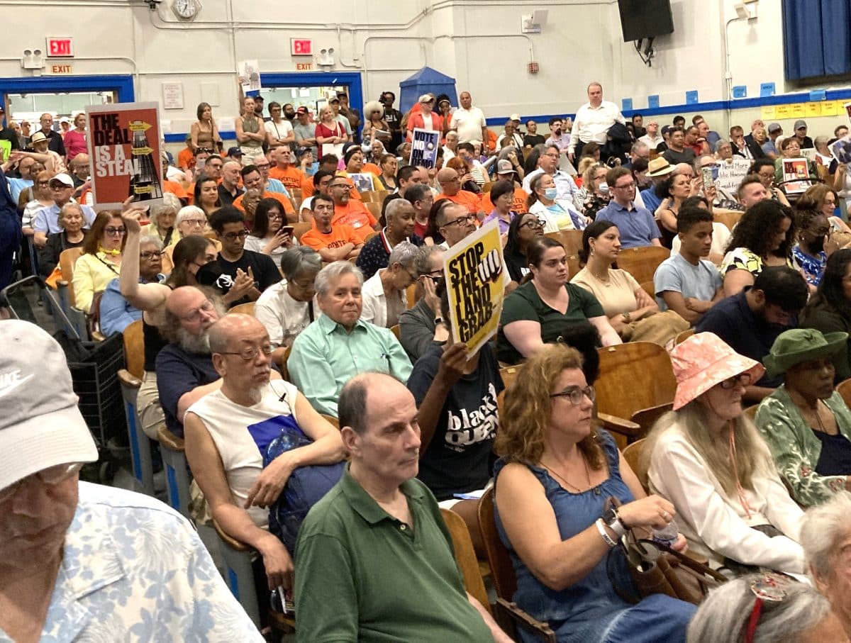 A large municipal room is packed with a diverse crowd of serious-looking people, most seated but with many standing at the back and filling two wide doorways at the back of the room. Several of the seated ones are holding posters. The three that are readable say "Stop the land grab," The deal is a steal," and "Vote No on Demo."