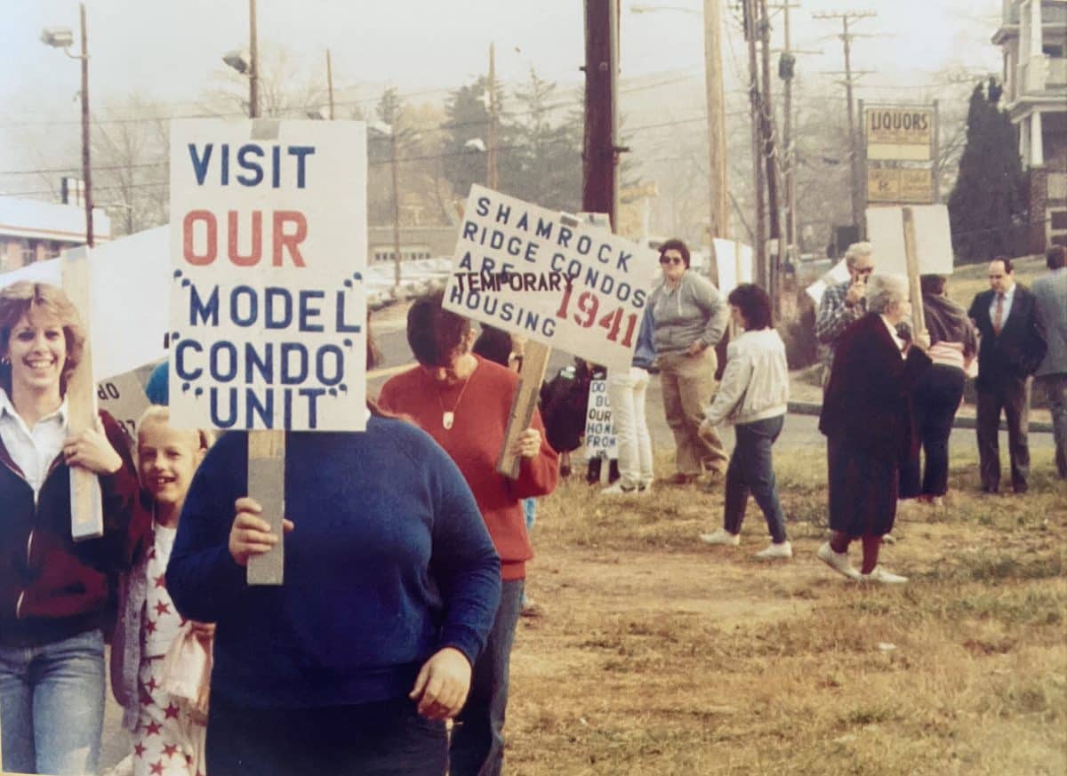 A 1980s scene of people picketing on a grassy roadside area. Visible signs say "Visit our Model Condo Unit," and "Shamrock Ridge Condos are Temporary Housing" (with 1941 in large red letters). The people picketing range in age from children to gray-hairs.
