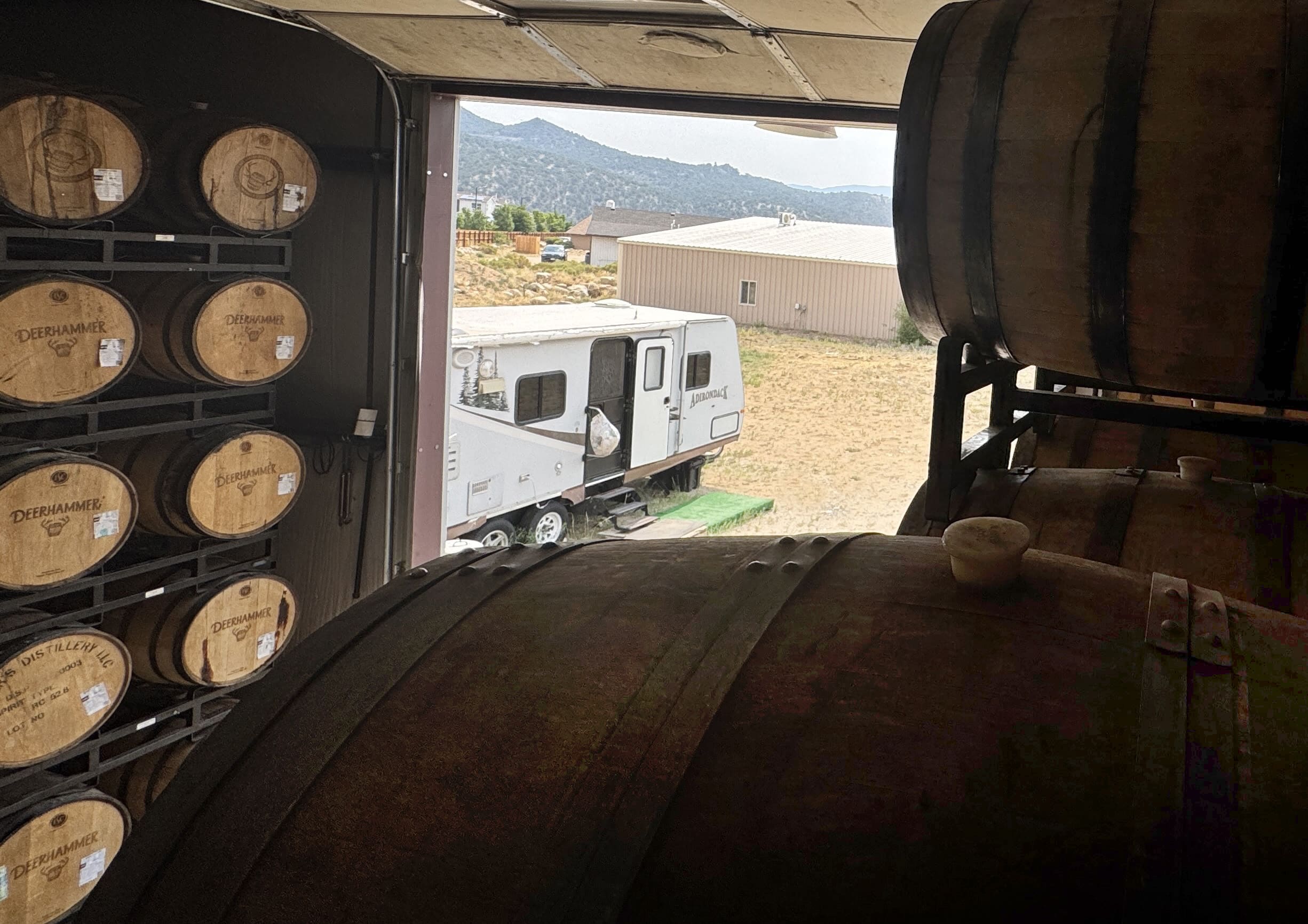 View from inside a distillery. In the foreground are large barrels, and against the wall at left are metal shelves holding smaller barrels, each with Deerhammer distillery name or logo printed on them. Outside the open garage-type door is a white camper van. Beyond it, some nondescript buildings and in the distance, mountains.