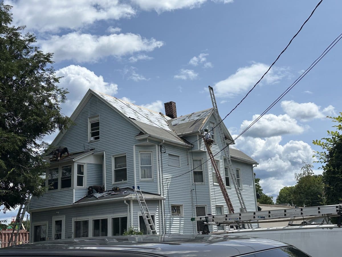 Under a bright sky with fluffy clouds, a large, three-story clapboard house with peaked roof and a gable on the side. Four ladders are propped against the house, leading to work being done on the roof and gutters. Workers can be seen on one ladder and a projecting roof over a second-floor sunroom. To the left is a tall deciduous tree.