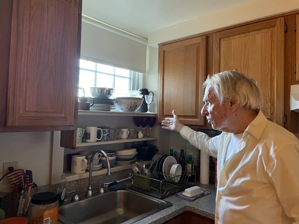 A man with white hair and a white shirt gestures toward a sink with mugs and dishes stacked behind.