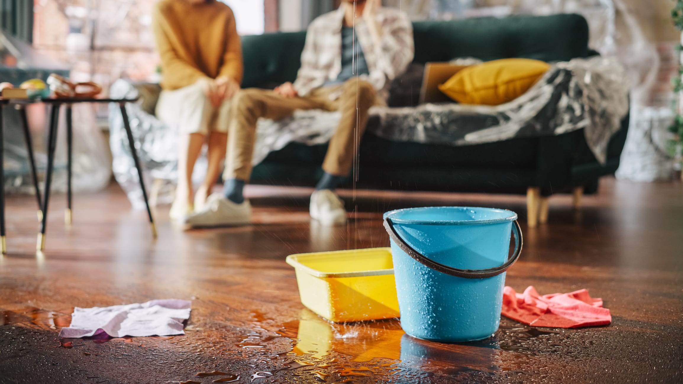 A leak pours into a bin and a bucket, on top of a wet hardwood floor. A couple sits on a couch in the background.