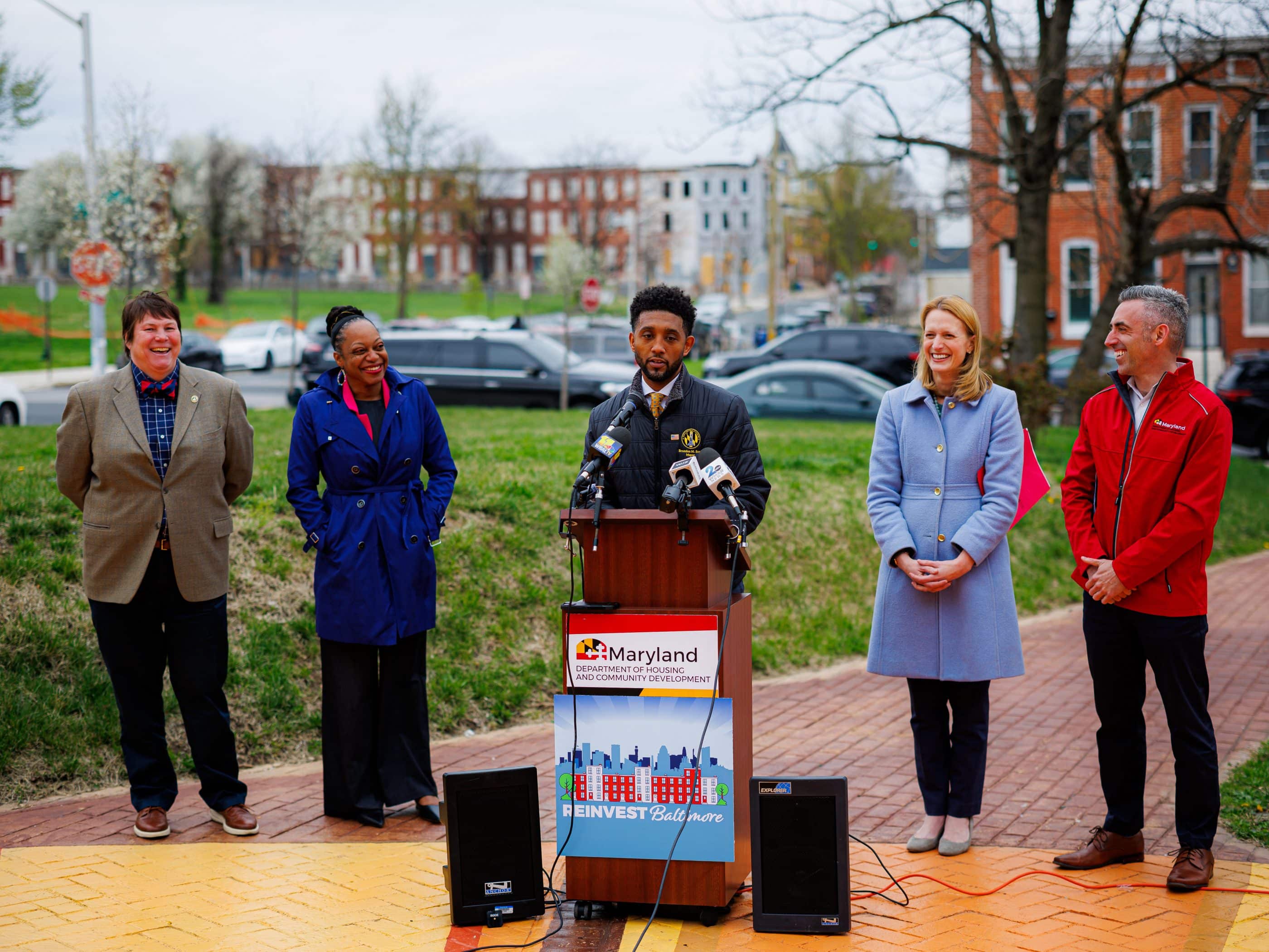 A group of five people stand outside during a press conference. Four people are smiling and looking at a Black man who speaks at a podium.