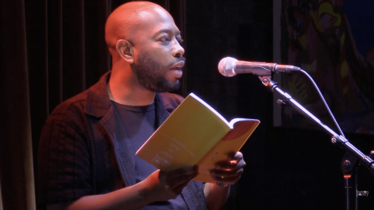 A Black man in a dark T-shirt and blazer recites poems from a yellow book into a microphone. The book says “Button Poetry” on the back cover.