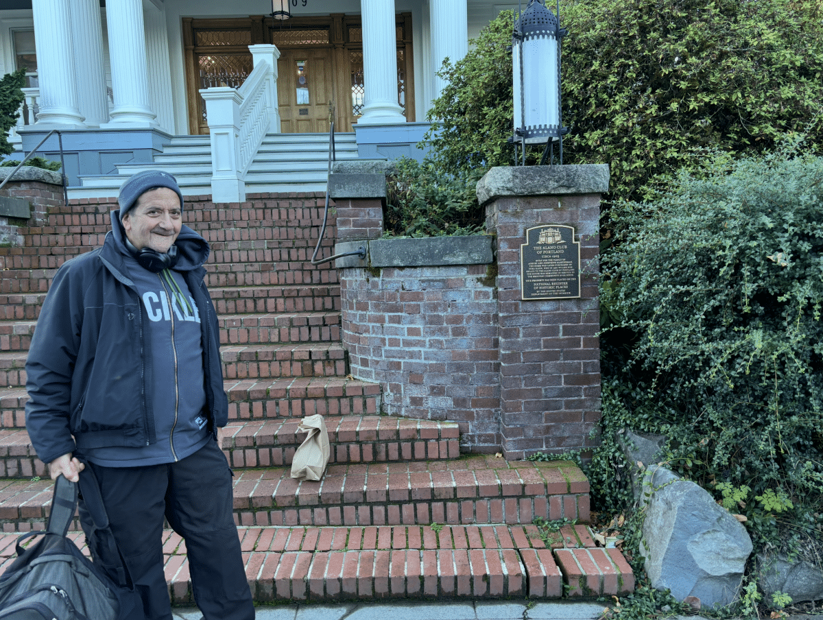 A middle-aged white man holding a gray and black backpack and wearing a black jacket, black pants, a gray hat, and headphones around his neck stands at the bottom of a brick staircase leading to a large historic building. A sign on the staircase reads, in part, "The Alano Club of Portland, circa 1905."