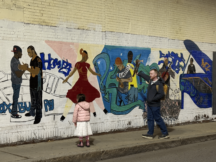A young girl in a pink jacket, white skirt, and pink boots (left) and a white man wearing a dark blue jacket, navy pants, and a yellow backpack (right) stand on a sidewalk in front of a colorful mural depicting different scenes of Black men and women playing instruments, shaking hands, and dancing.