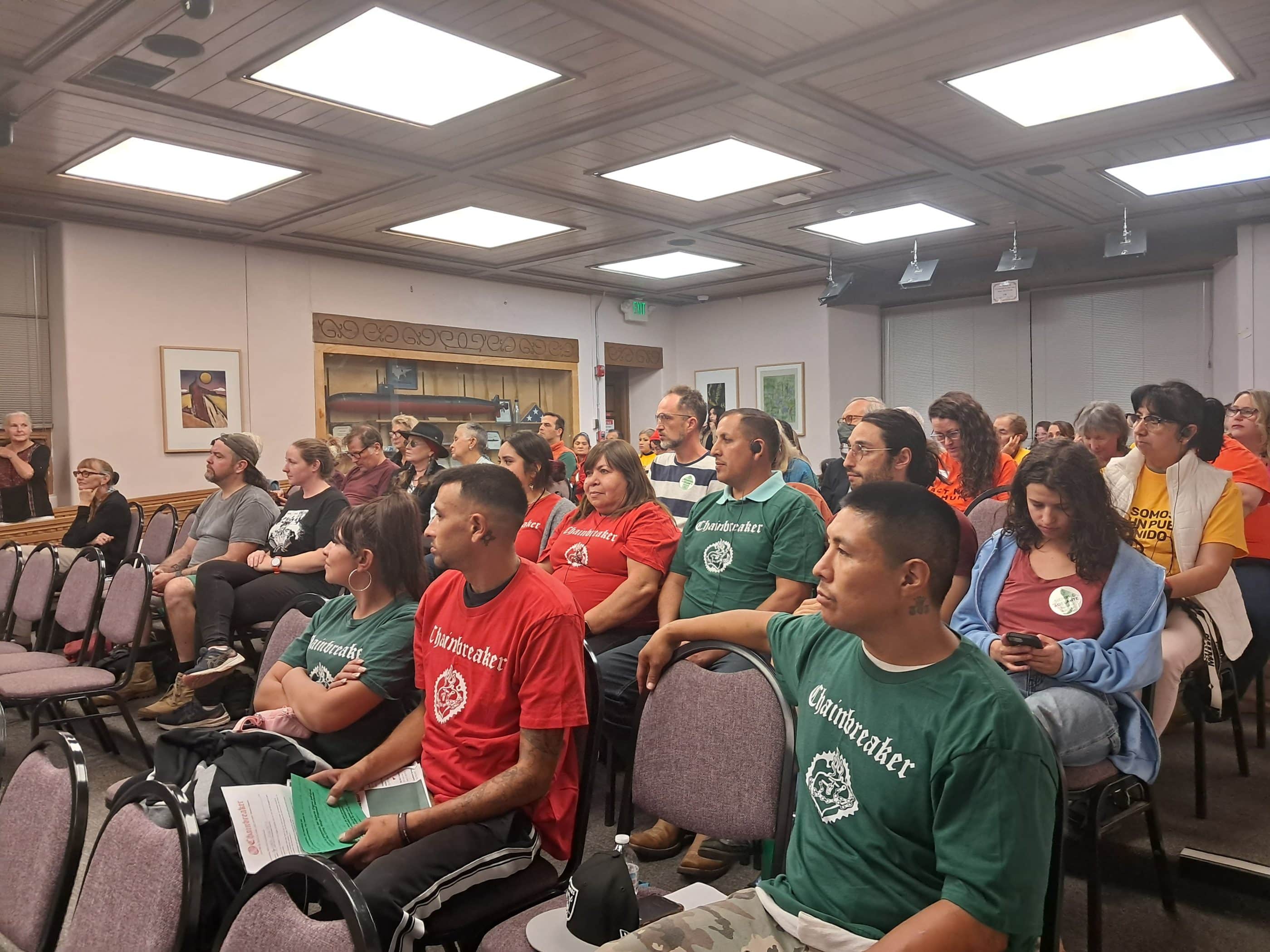 People of various ages, races, and ethnicities sitting on chairs in what appears to be a meeting room. Many of the people are wearing green or red T-shirts that say "Chainbreaker" in white text on the upper-chest area.
