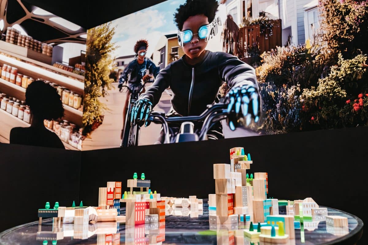 A group of building blocks on a table with buildings printed on them. Above them is a projected image of two boys on bikes wearing futuristic gear.