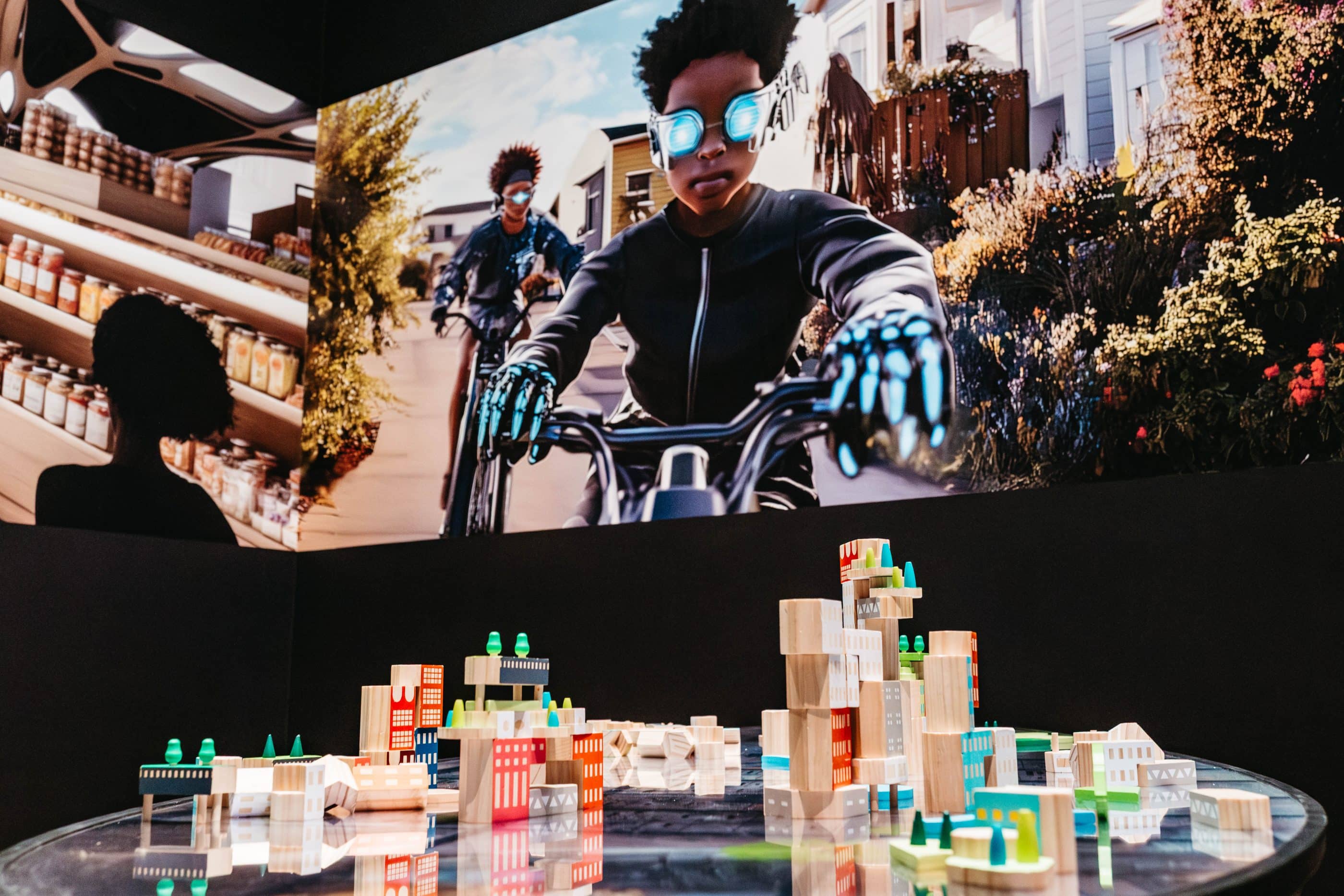 A group of building blocks on a table with buildings printed on them. Above them is a projected image of two boys on bikes wearing futuristic gear.