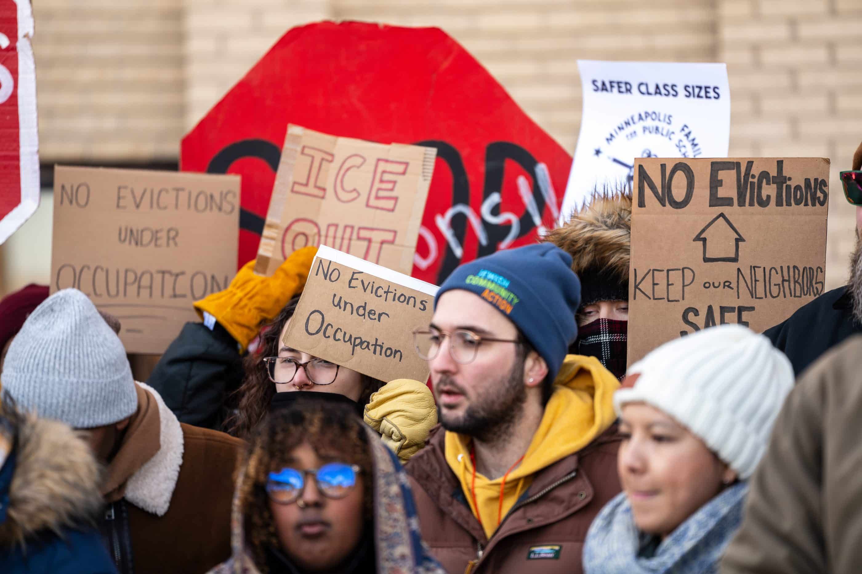 A group of people of different ages, genders, and races stands outside, holding up cardboard signs that say things like "ICE OUT" and "No evictions under occupation." Many of the people are bundled up in jackets and beanies. A beige brick wall appears in the background.