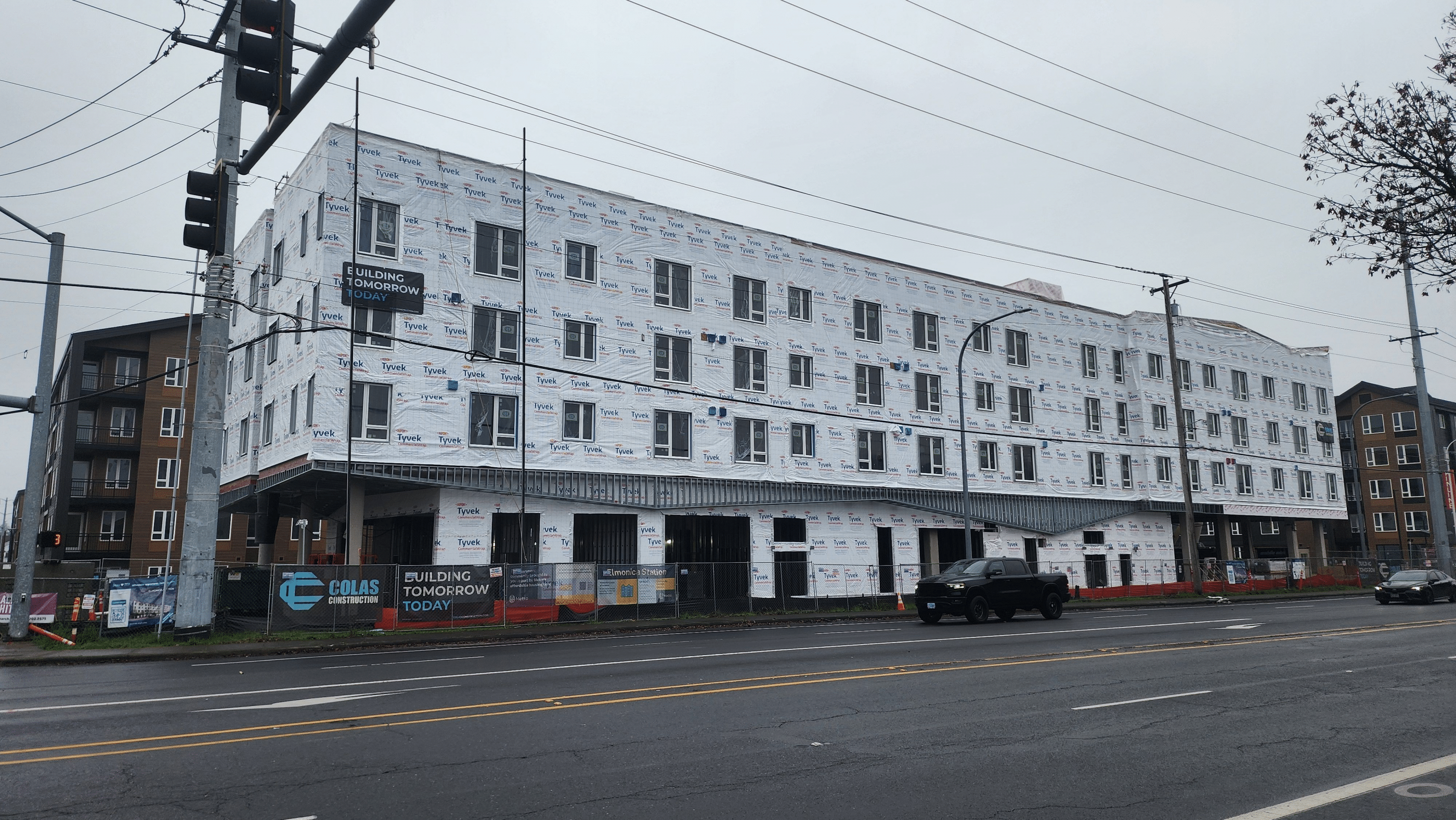 A large housing development under construction. The building is covered in Tyvek wrap and sits behind fencing with blue and white signs that read "Colas Construction: Building Tomorrow Today."