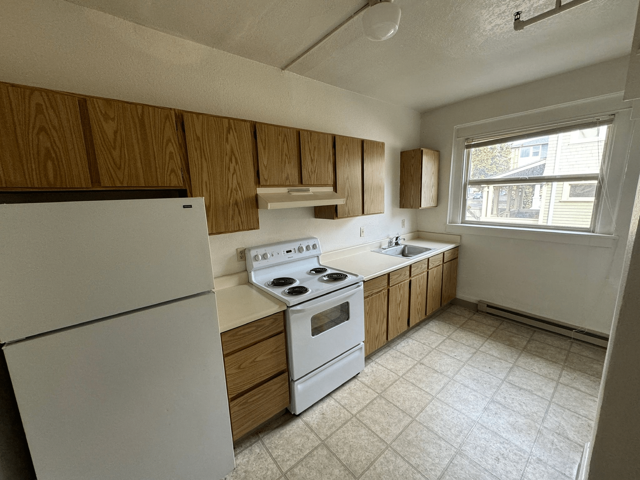 A simple kitchen in an apartment building, featuring a white fridge, white four-burner electric stove, brown laminate cabinets, and beige tiled flooring. A large kitchen window looks out on a tree-lined street with residential homes.