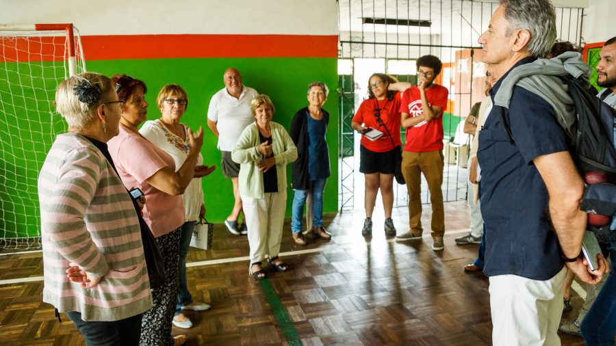 A group of men and women standing in a circle in a gymnasium or community recreation room. The people appear to be holding a community meeting.