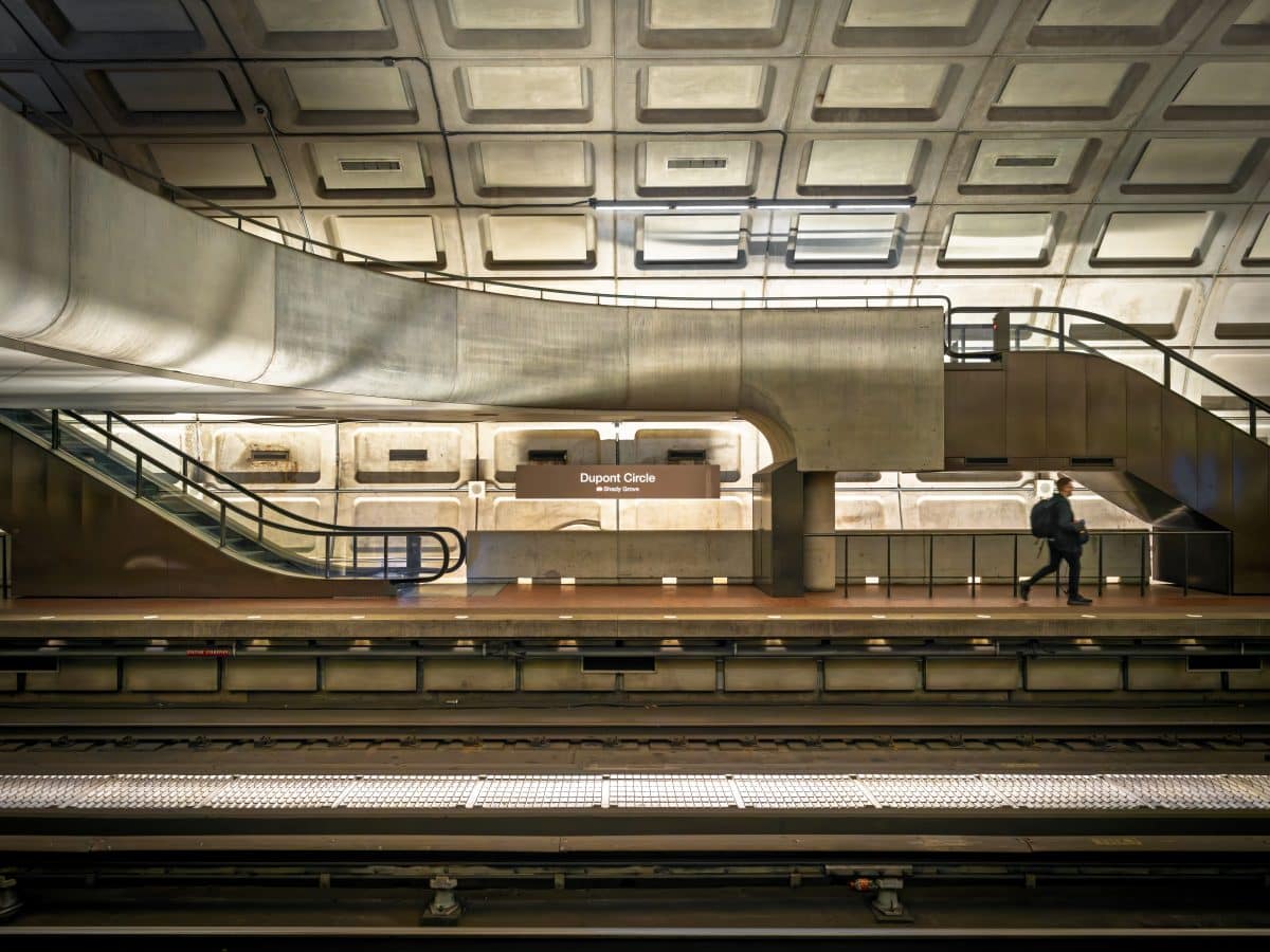 The interior of Dupont Circle train station in Washington, D.C., showing a train track, train platform, and Brutalist-style architecture, including large staircases and walking paths. A white man wearing dark clothing and a large backpack crosses the train platform in the right-hand corner.