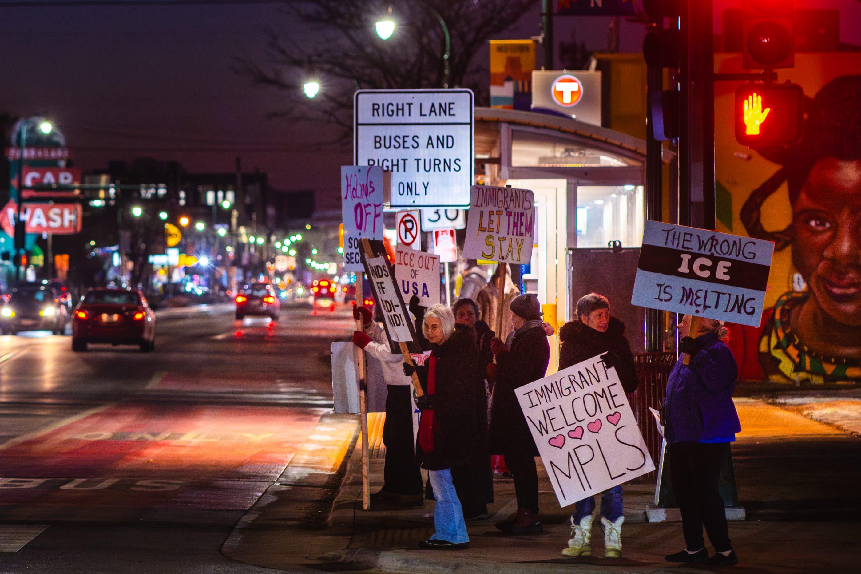 A group of older women stand on a sidewalk next to a busy street in Minneapolis. The women hold protest signs that say things like "The wrong ICE is melting," "ICE out of USA," and "Immigrants welcome MPLS."