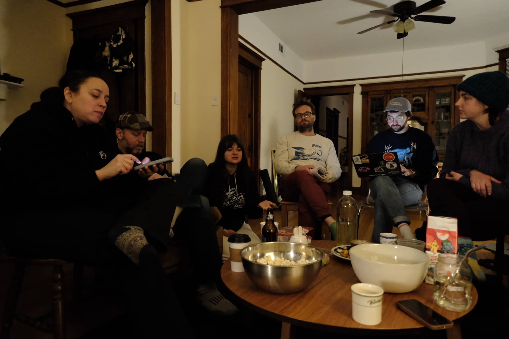 Six white, mostly young, people sitting in chairs around a round coffee table. One person has a laptop on their lap, and another is on their phone. They appear to be holding a meeting.